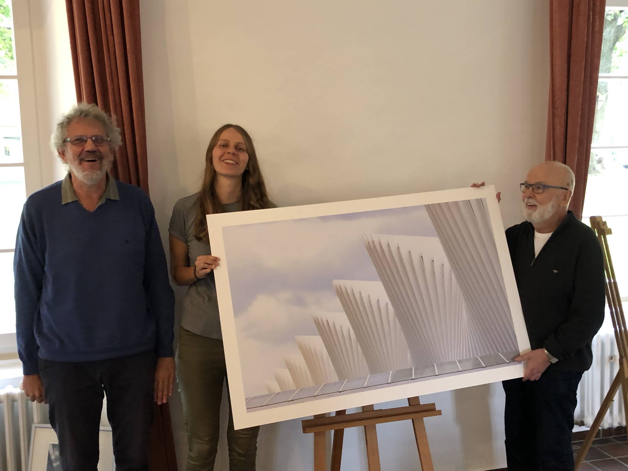  Detlev und Larissa Kahler präsentieren gemeinsam mit Ausstellungsmacher Roland Brozio eine ganz besondere Arbeit von Sylvia Kahler: Es zeigt einen Bahnhof in Reggio Emilia und wurde von der Jüchener Künstlerin im Jahre 2017 beim „Siena International Photo Award“ eingereicht. „Sie kam damals in die Reihe der besonders Ausgezeichneten. Die Verleihungszeremonie ist noch heute als Video zu finden“, strahlt der Witwer, der anfängt: „Sie war soviel gebildeter als ich.“ 
  
