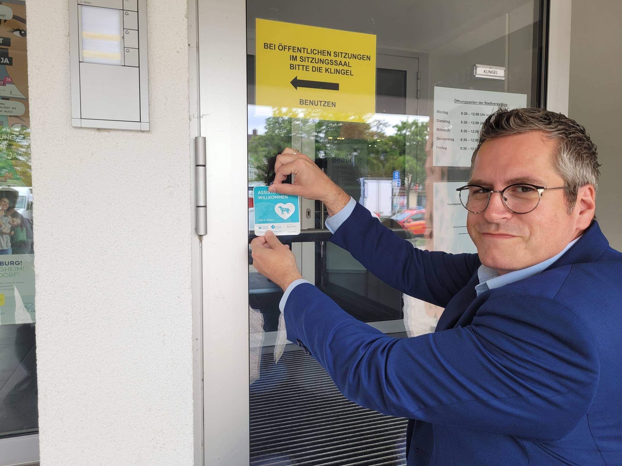 „Assistenzhunde willkommen“: Bürgermeister Sascha Solbach bringt den passenden Aufkleber am Rathaus an.